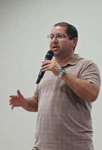 Um homem de pele clara, cabelo castanho escuro e óculos, trajando uma camisa polo bege com um padrão xadrez discreto, fala em um microfone. Ele usa um relógio de pulso prateado no braço esquerdo e gesticula com as duas mãos. O fundo é uma parede branca lisa.