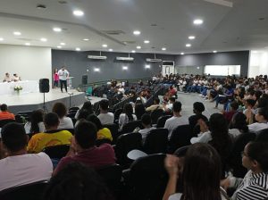 A imagem mostra uma plateia sentada em um auditório, de frente para um palco onde várias pessoas estão presentes. Uma pessoa no palco está falando ativamente, gesticulando em direção à plateia, enquanto outras estão sentadas a uma mesa. A plateia é composta por um grupo diverso de pessoas, parecendo ter várias idades, embora muitos pareçam ser jovens adultos ou estudantes.