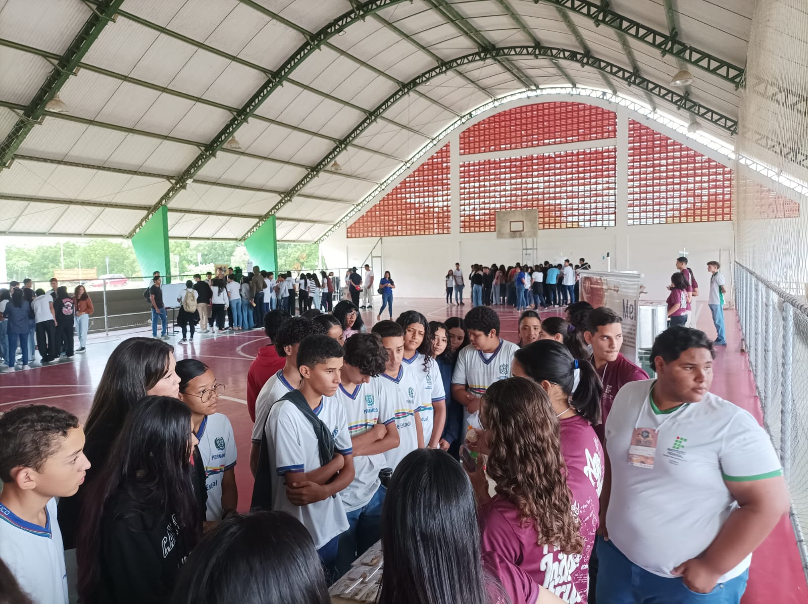 Um grupo de estudantes está reunido na quadra do campus Ouricuri. Eles são diversos em aparência, com várias cores e estilos de cabelo, e a maioria está vestindo roupas casuais, algumas com camisetas que podem fazer parte de um uniforme escolar ou camiseta de evento. O ambiente sugere um evento como uma feira escolar, exposição ou um dia aberto, já que há muitas pessoas interagindo e algumas exibições ou estandes visíveis ao fundo.