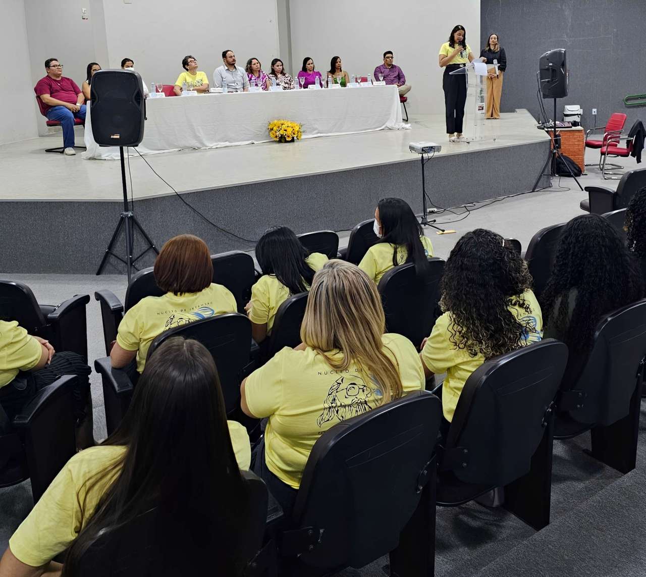Auditório com plateia formada majoritariamente por mulheres usando camisetas amarelas com ilustração de Paulo Freire nas costas, sentadas de frente para um palco. No palco, há uma mesa composta por autoridades e convidados, com toalha branca e arranjo de flores amarelas ao centro. Duas mulheres estão em pé à direita, uma delas discursando ao microfone. Ao fundo, paredes claras e ambiente bem iluminado. Cena retrata a cerimônia de certificação do curso de Formação e Aperfeiçoamento em Práticas Pedagógicas na Perspectiva de Paulo Freire, realizada no campus Ouricuri do IFSertãoPE.