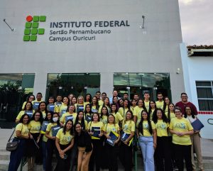 Um grupo grande de cerca de 40 pessoas, a maioria mulheres, está em frente a um prédio cinza com janelas espelhadas. Todos estão usando camisetas amarelas com um design azul e verde na frente. Eles estão olhando para a câmera e sorrindo. O prédio tem o logotipo do Instituto Federal, um quadrado verde composto por quadrados menores, e o texto "INSTITUTO FEDERAL Sertão Pernambucano Campus Ouricuri" em letras grandes e pretas acima da entrada. O céu está um pouco nublado.