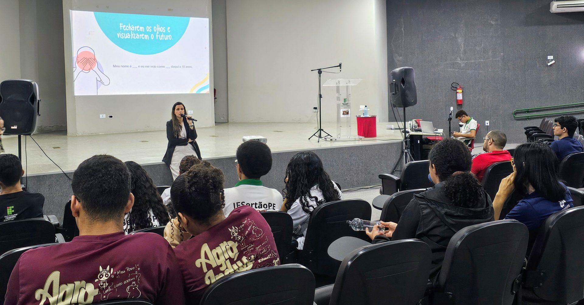 Auditório com estudantes sentados assistindo a uma palestra. À frente, uma mulher, identificada como a psicóloga Paula Leal, está de pé com um microfone na mão, falando ao público. No fundo, há um telão com uma projeção que diz: “Fecharem os olhos e visualizarem o futuro. Meu nome é ___ e eu me vejo como ___ daqui a 10 anos.” A palestrante está vestida com blazer escuro e calça clara. À direita, há um púlpito de acrílico com o logotipo do IFSertãoPE e equipamentos de som. Os estudantes usam camisas do Instituto Federal e de cursos técnicos.