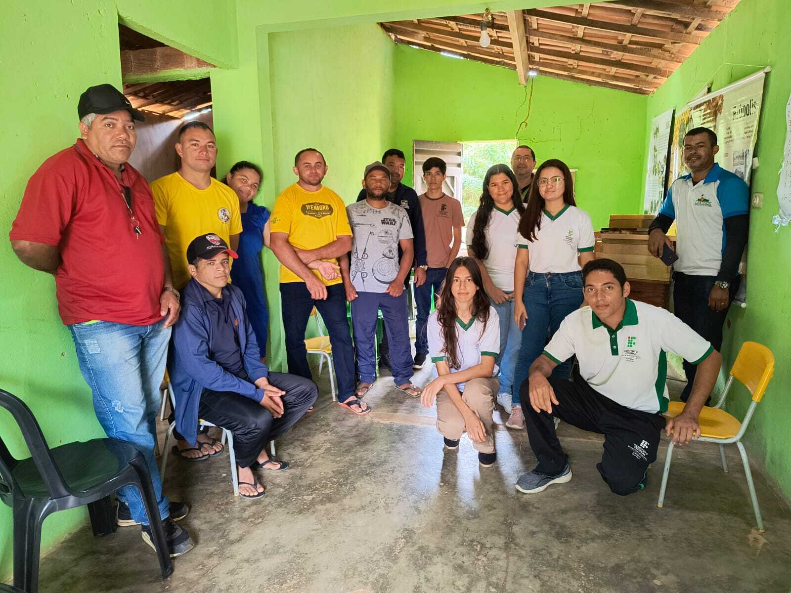 A imagem mostra um grupo de 15 pessoas, homens e mulheres, de diferentes idades, reunidas em uma sala com paredes verde-claras. O ambiente parece rústico, com telhas e parte da estrutura do telhado visíveis. Da esquerda para a direita, em pé, há um homem de camisa vermelha e boné preto, seguido por outro de camisa amarela. Ao fundo, um homem de camisa azul escura. Mais ao centro, um homem de camisa amarela e outro de camiseta cinza clara com uma estampa. À direita deles, um jovem de camiseta escura e boné. Mais ao centro da imagem, um homem de camisa azul-claro, uma mulher de blusa branca e calça jeans, e outra mulher de blusa branca com um logotipo verde. À extrema direita, em pé, um homem de camisa polo branca e azul com um logotipo. Agachados ou sentados, da esquerda para a direita, há um homem sentado em uma cadeira escura, um homem sentado no chão de boné, uma mulher agachada no centro da foto, e um jovem de camisa polo branca e verde com um logotipo, agachado em primeiro plano. Há cadeiras amarelas no canto inferior direito e algumas caixas de madeira empilhadas ao fundo. Na parede do fundo, à direita, é possível ver alguns pôsteres ou gráficos fixados.