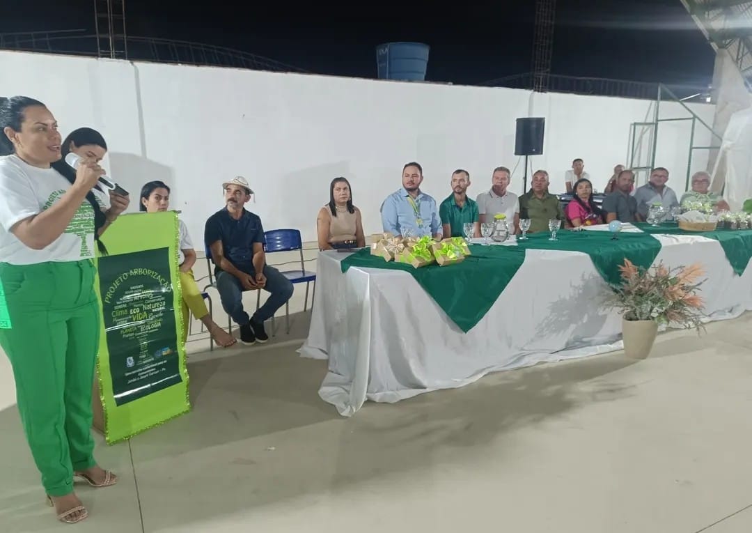 Uma tomada completa mostra um evento noturno ao ar livre com uma mulher no lado esquerdo do quadro falando em um microfone. Ela tem cabelos escuros, está vestindo uma camiseta branca com um desenho de árvore verde e calças verdes. À sua direita, outra mulher está segurando uma placa verde com texto. No centro e no lado direito da imagem, uma longa mesa está montada com uma toalha de mesa branca e um caminho de mesa verde. Atrás da mesa, várias pessoas estão sentadas, observando a oradora. Elas parecem ser autoridades ou convidados de honra. O fundo consiste em uma parede branca e algumas árvores, e a área é iluminada por luzes artificiais.