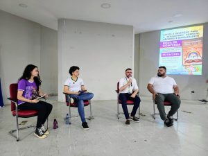  Uma imagem em close de quatro pessoas sentadas em cadeiras dispostas em semicírculo, de frente para a câmera, com um púlpito e um monitor atrás delas. As duas pessoas mais à esquerda são mulheres, e as duas mais à direita são homens. A mulher mais à esquerda veste uma camisa roxa e calça preta. A mulher sentada ao lado dela veste uma camisa branca e calça jeans. Ela está segurando um caderno e uma caneta na mão esquerda. O homem sentado ao lado dela veste uma camisa social branca e calça azul escura, e está usando um microfone na mão direita. O homem mais à direita veste uma camisa branca e calça verde escura. Todos os quatro estão usando tênis. O fundo da imagem é uma parede branca e lisa, com uma projeção em um telão branco à direita. O telão exibe informações sobre uma "RODA DE CONVERSA" com o tema "AFETOS E EDUCAÇÃO PARA A SEXUALIDADE: CORPO(S), GÊNERO(S) E SEXUALIDADE(S)". Há também a data "25/06/2025" e o local "auditório do Campus Ouricuri". Uma garrafa de água pode ser vista no chão entre as duas mulheres.