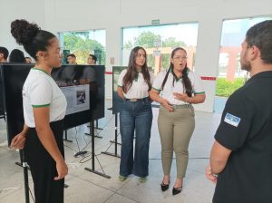 Três estudantes, usando camisetas brancas com detalhes verdes, apresentam um projeto pedagógico em uma tela digital. Duas delas, no centro, conversam com um homem que as ouve, enquanto a terceira observa a tela. A cena ocorre no pátio do Campus Ouricuri, durante a Mostra Pedagógicas de Projetos