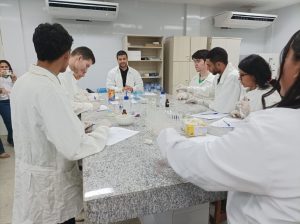 Um grupo de seis estudantes, todos usando jalecos brancos e luvas de proteção, está reunido em volta de uma bancada de laboratório de granito. Eles estão a manusear tubos de ensaio, frascos de vidro e papéis, enquanto um dos jovens no centro conversa com o grupo. A cena acontece num laboratório de química, com armários e equipamentos visíveis ao fundo.