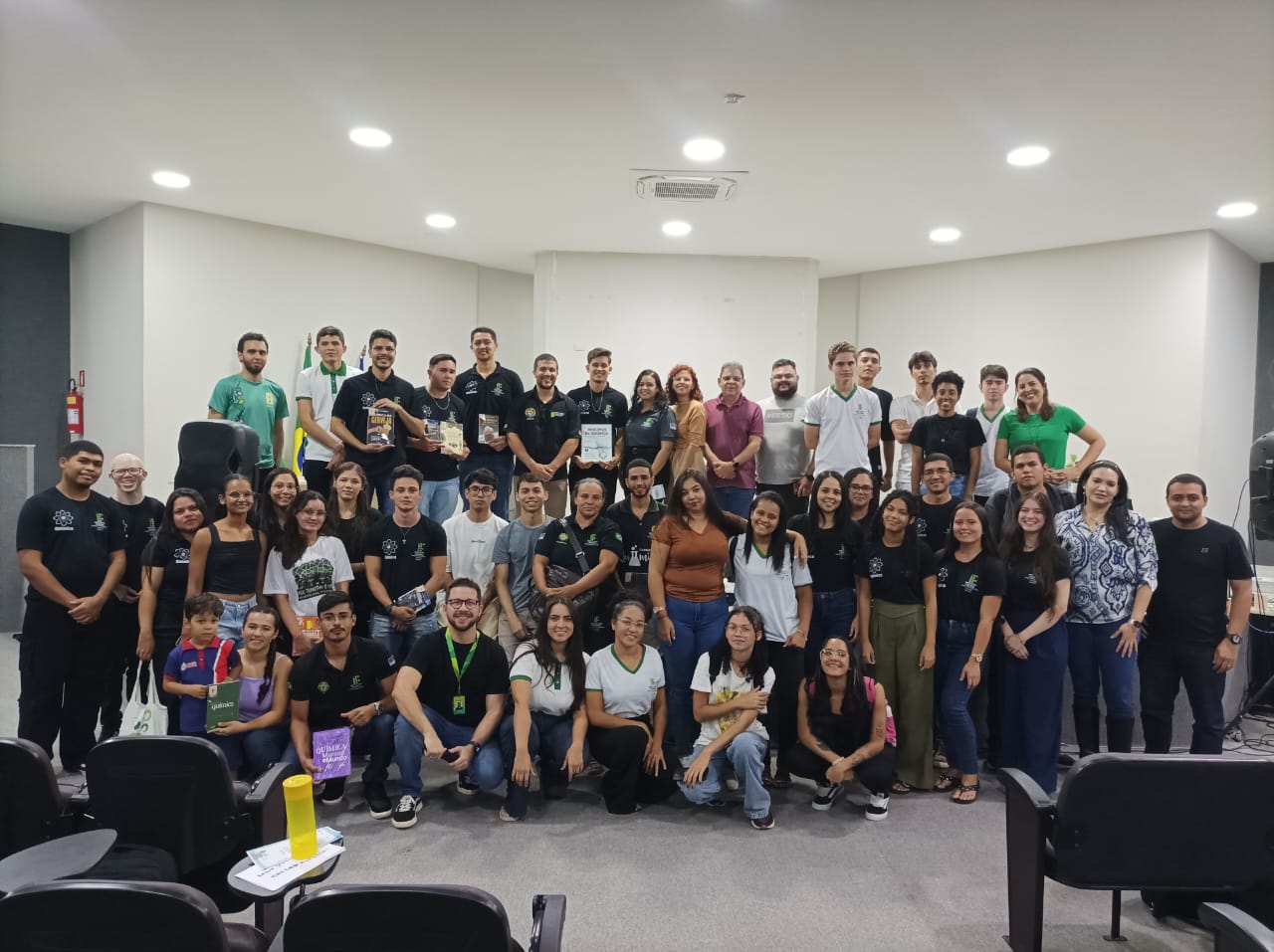 Uma foto de grupo grande tirada num auditório. Cerca de 40 pessoas de idades e géneros variados estão de pé e sentadas, posando para a câmara. A maioria veste camisolas pretas com logótipos brancos, e algumas pessoas no centro seguram livros ou prémios. No lado esquerdo, são visíveis as bandeiras do Brasil e de Pernambuco.