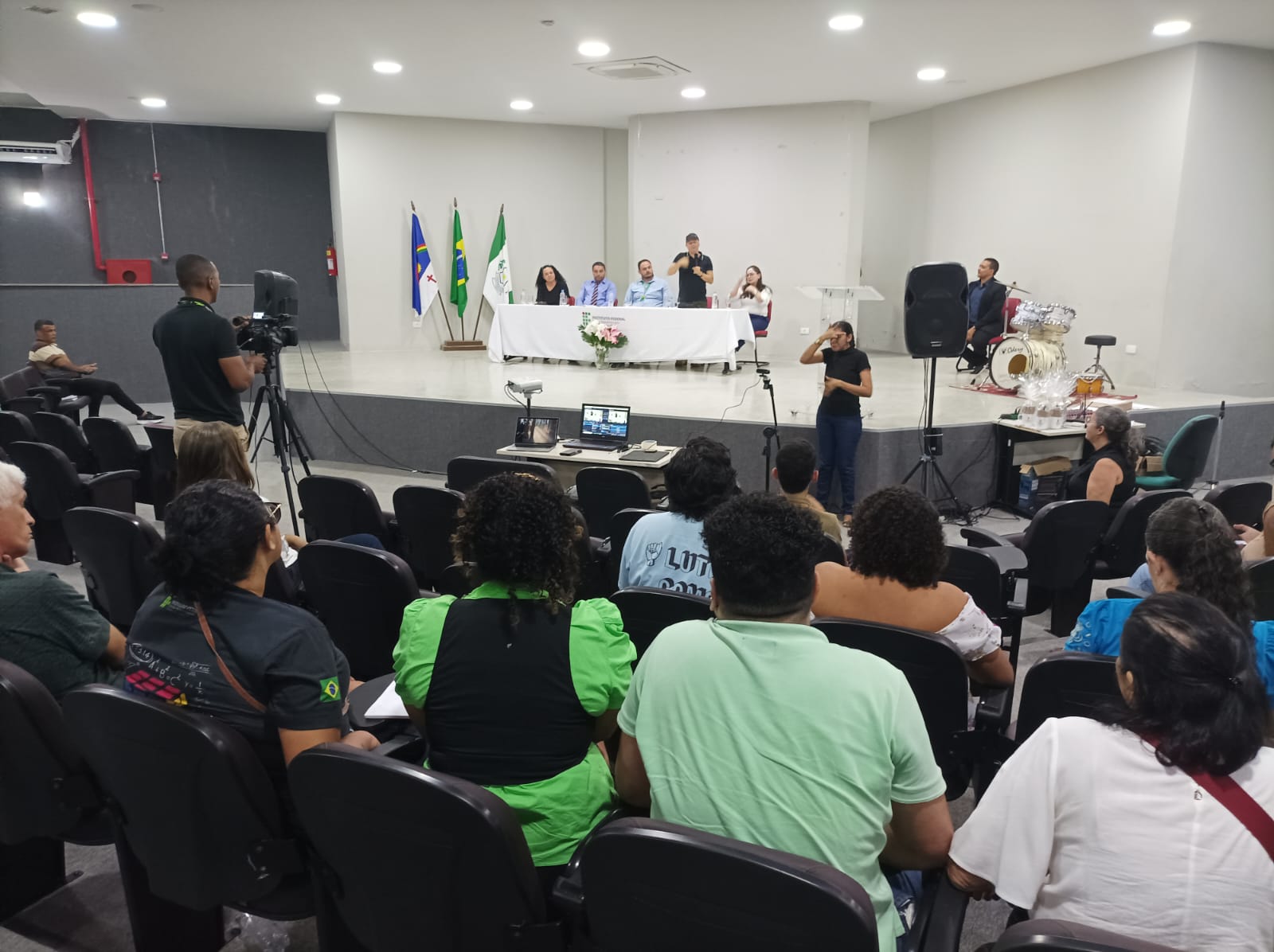 A imagem mostra uma palestra em um auditório. No palco, há uma mesa com seis pessoas sentadas, e atrás delas estão três bandeiras. Há uma pessoa de pé no palco, que parece estar fazendo uma apresentação, possivelmente em Libras. Próximo a essa pessoa, há uma banda com um baterista. O público está sentado em cadeiras pretas, de costas para a câmera, assistindo à palestra. Há um cinegrafista em pé, filmando o evento, e um técnico operando um notebook e outros equipamentos em uma mesa pequena no centro do auditório.