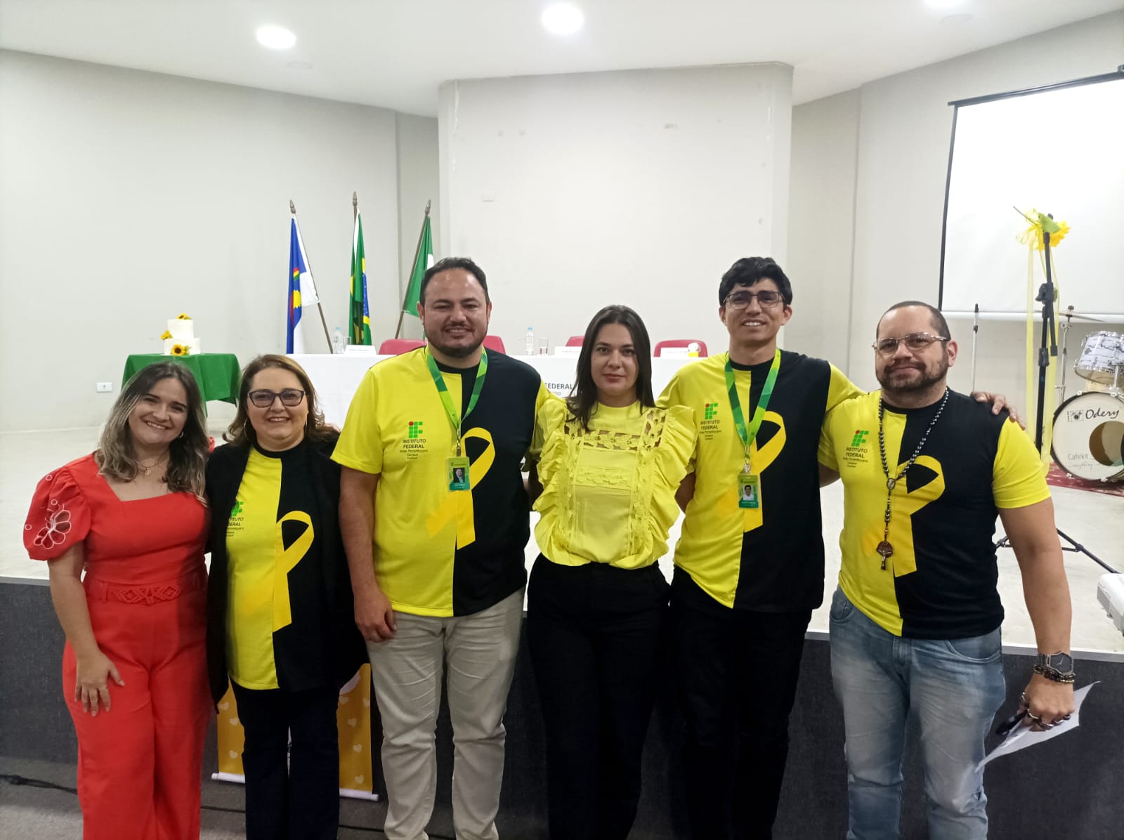 A imagem é uma fotografia de um grupo de seis pessoas, quatro homens e duas mulheres, posando juntas em um palco ou auditório decorado para a campanha Setembro Amarelo. Descrição do Grupo e Vestimentas: Quatro das pessoas (três homens e uma mulher) vestem camisetas que combinam as cores preto e amarelo, com um detalhe gráfico que remete ao laço do Setembro Amarelo ou a um logotipo do Instituto Federal (IF). Uma mulher à esquerda veste um conjunto de calça e blusa de manga bufante na cor vermelha alaranjada. Uma segunda mulher, ao lado da de vermelho, veste uma camiseta preta com detalhes em amarelo e o laço. Os homens vestem camisetas pretas e amarelas combinadas com calças em tons claros (cáqui ou jeans). Fundo da Cena: O grupo está em pé em um ambiente interno que parece ser um palco ou auditório. Ao fundo, vê-se: Uma mesa com toalha branca. Bandeiras (provavelmente do Brasil, de Pernambuco ou do IF). Um pequeno bolo de aniversário ou celebração. À direita, há um telão (parcialmente visível), um pedestal de microfone e equipamentos de som, incluindo um bumbo de bateria com a marca Odery, decorados com fitas amarelas.