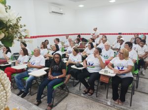 Sala de aula cheia, com pessoas da terceira idade sentadas em carteiras escolares. A maioria veste camisetas brancas do projeto, enquanto ouvem atentos à programação da solenidade.