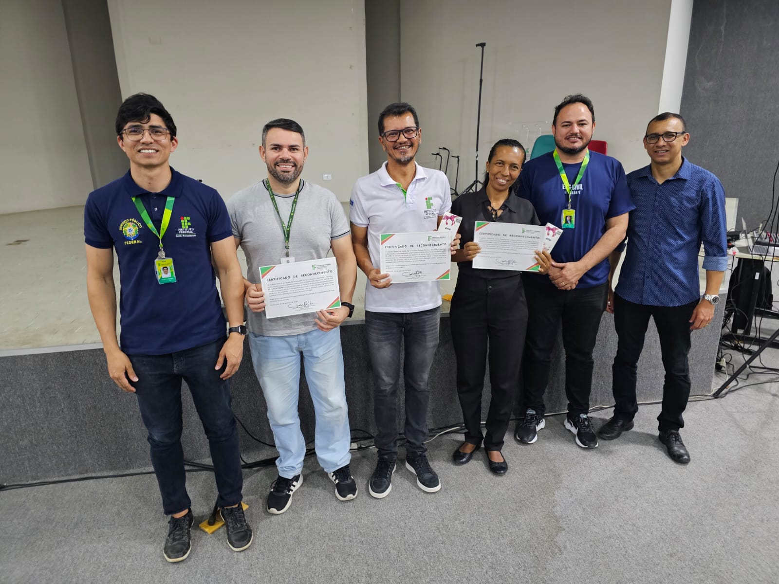 Uma fotografia tirada durante o evento da campanha institucional “Valorizamos quem faz a diferença: o Assistente de Aluno” mostra seis pessoas de pé, alinhadas em uma sala. Três homens à esquerda usam camisetas cinza ou azul-marinho. Duas das pessoas no meio, um homem e uma mulher, vestem camisetas polo brancas ou blusas pretas. Um homem à direita usa uma camisa azul com estampa. A maioria segura certificados ou pequenos brindes, sendo estes os assistentes de alunos que estão sendo homenageados. O fundo da sala tem paredes cinzas, uma pequena mesa preta e equipamentos eletrônicos.