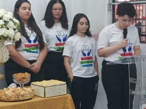 Quatro jovens alunas monitoras do projeto estão em frente à mesa decorada, todas com camisetas brancas do evento. Uma delas lê um texto no púlpito com microfone, enquanto as outras acompanham atentas.