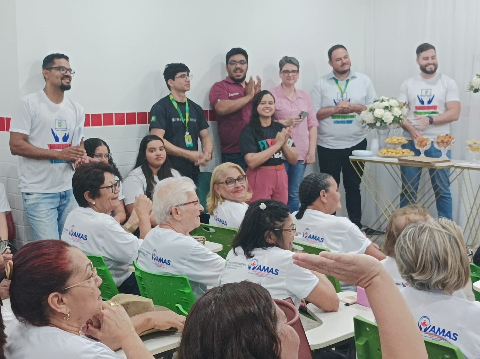 Grupo de organizadores e convidados em pé, de frente para a plateia, aplaudindo durante a cerimônia. À frente, idosos sentados com camisetas do projeto e da AMAS. Ao lado, mesa decorada com flores e salgados.