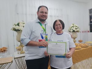 Um homem e uma mulher posam sorrindo para a câmera. Ambos vestem camisetas brancas do projeto Inclusão Digital e Internet Segura para a Terceira Idade. O homem segura um certificado em nome da mulher. Ao fundo, mesa decorada com flores e salgados.