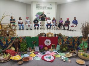 Esta fotografia em plano médio captura o palco e a exposição de produtos durante a realização do seminário.Mesa de Autoridades: No palco, cerca de dez pessoas (mulheres e homens), vestidas de forma casual, estão sentadas em cadeiras dispostas em uma linha. Um homem em pé, no centro, está falando ao microfone. Decoração e Exposição: A frente do palco é decorada com uma toalha floral e uma exposição rica em cores e texturas, simbolizando a agricultura familiar. São visíveis: bananas, batata-doce, abóboras, milho, cocos, raízes, pimentas, ovos, e sementes em cestos. Também há livros, cadernos e folhetos do Movimento dos Pequenos Agricultores (MPA) e um estandarte com a bandeira do MPA. Banner: Acima, um banner branco exibe o título: "Seminário Estadual de Agroecologia e Abastecimento Popular" e "I Encontro de Guardiãs da Agrobiodiversidade", com as datas 03 e 04 de Outubro.