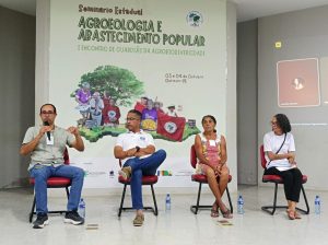 Fotografia frontal de uma mesa de diálogo durante o seminário.Integrantes da Mesa: Quatro pessoas estão sentadas em cadeiras no palco. O primeiro, à esquerda, fala ao microfone, gesticulando. Os demais (um homem e duas mulheres) ouvem. Projeção/Fundo: O banner do seminário, com o título e ilustrações de agricultoras, serve de fundo para a mesa. No lado direito, um quadro branco projetado mostra participantes online, incluindo "Joselita Tavares".