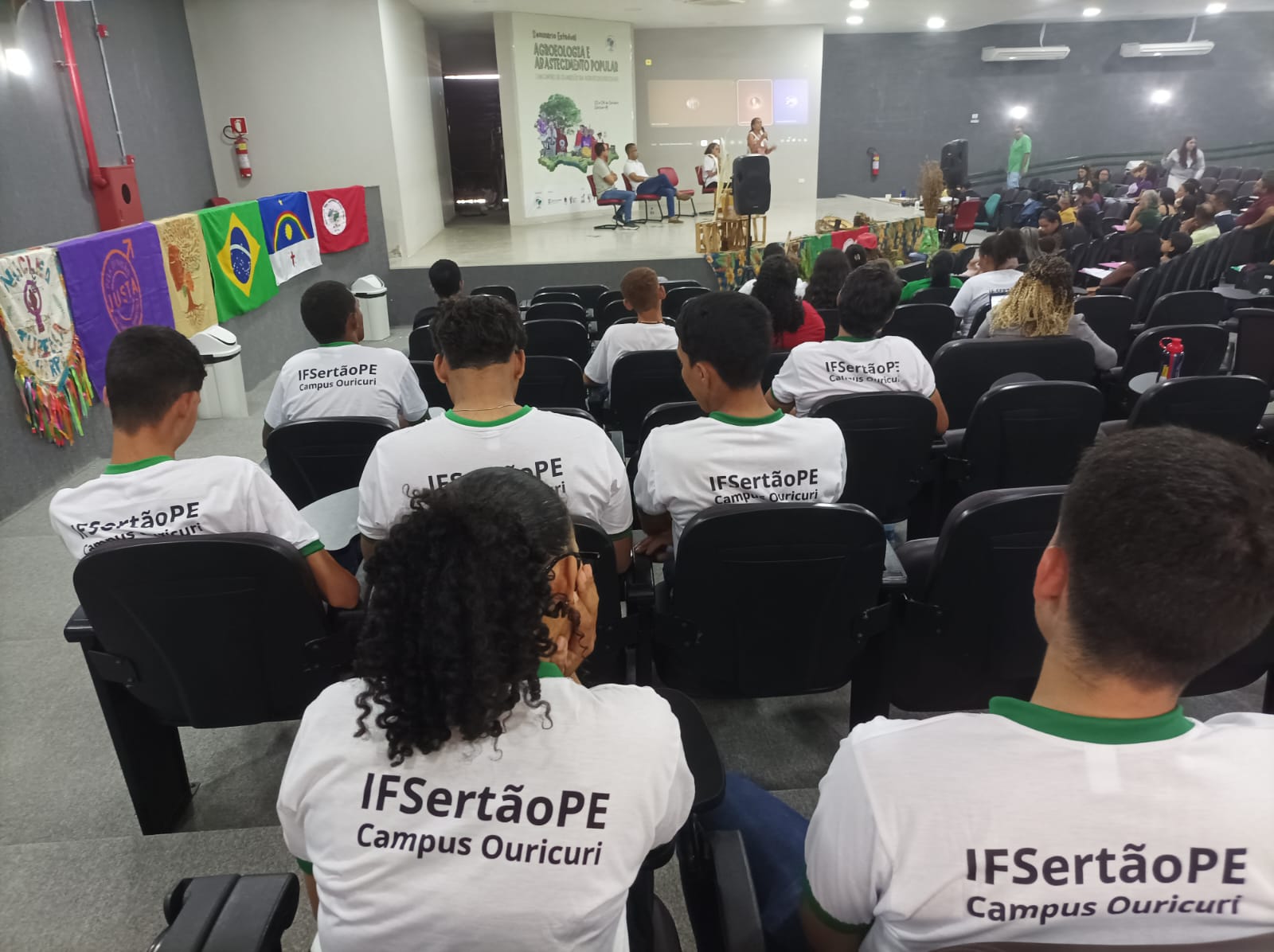 A imagem é uma foto panorâmica de um evento sendo realizado no auditório do Campus Ouricuri. Público: A foto é tirada da parte de trás da plateia, focando nas nucas e costas de vários estudantes sentados em cadeiras escuras. Muitos vestem camisetas brancas com a inscrição "IFSertãoPE Campus Ouricuri" nas costas. Cenário: Ao fundo, está o palco, onde algumas pessoas estão sentadas à mesa (mesa de honra) e uma oradora está em pé, ao lado de um púlpito. Acima, um banner ou projeção exibe o título: "Seminário Estadual de Agroecologia e Abastecimento Popular". Decoração: Na lateral esquerda do auditório, bandeiras e tecidos coloridos estão pendurados, incluindo a bandeira do Brasil, a de Pernambuco e a do IFSertãoPE. A imagem sugere a realização do Seminário Estadual de Agroecologia e do Encontro de Guardiãs da Agrobiodiversidade.