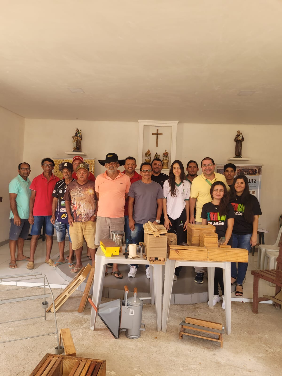 Foto de grupo tirada na Igreja dos Santos Reis, no bairro Várzea do Meio, em Bodocó – PE, durante o terceiro Dia de Campo do projeto de extensão 'NUPEA, Além dos Muros do Campus Ouricuri', realizado pelo Núcleo de Pesquisa e Extensão em Abelhas (NUPEA) do IFSertãoPE. O grupo é composto por membros do NUPEA (identificáveis pelas camisas com a logo da instituição ou do projeto) e produtores locais. Eles estão em pé, posando atrás e ao redor de uma mesa com diversos materiais e equipamentos de apicultura, como caixas de madeira para abelhas (colmeias), quadros internos e outros utensílios. Ao fundo, um altar simples com uma cruz e imagens de santos indica o local do evento.