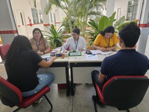 Equipe do Hemocentro de Pernambuco (Hemope) orienta servidores e estudantes do IFSertãoPE em uma mesa durante o evento Dia D de Saúde no Campus Ouricuri. Três profissionais do Hemope, identificadas com crachás, conversam e preenchem formulários enquanto duas pessoas são atendidas. Ao fundo, há plantas e o corredor do campus.