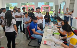 Profissionais do Centro de Testagem e Aconselhamento (CTA) realizam testes rápidos para HIV, sífilis e hepatites B e C durante o evento Dia D de Saúde no Campus Ouricuri do IFSertãoPE. Na imagem, três profissionais atendem um participante sentado à mesa, enquanto estudantes e colaboradores terceirizados aguardam na fila, observando o procedimento.