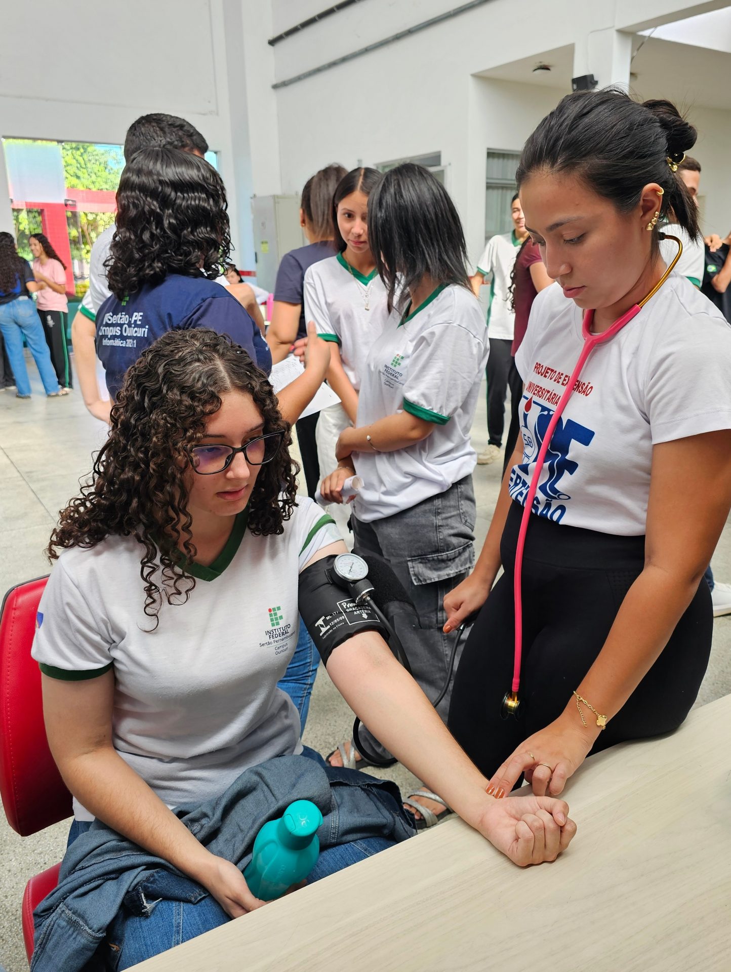 Em um espaço interno do campus, uma estudante com camiseta do IFSertãoPE está sentada enquanto outra mede sua pressão arterial com um esfigmomanômetro e estetoscópio. Ao fundo, outros estudantes conversam e aguardam atendimento durante o evento Dia D de Saúde no Campus Ouricuri.