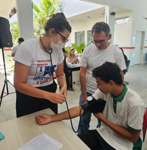 Uma estudante do curso de Enfermagem da Universidade de Pernambuco (UPE) – Campus Ouricuri – aferindo a pressão arterial de um aluno do Campus Ouricuri do Instituto Federal do Sertão Pernambucano (IFSertãoPE) durante o evento Dia D de Saúde. Ao lado, um professor Elias Coelho do IFSertãoPE observa o procedimento, realizado em um espaço interno do campus.