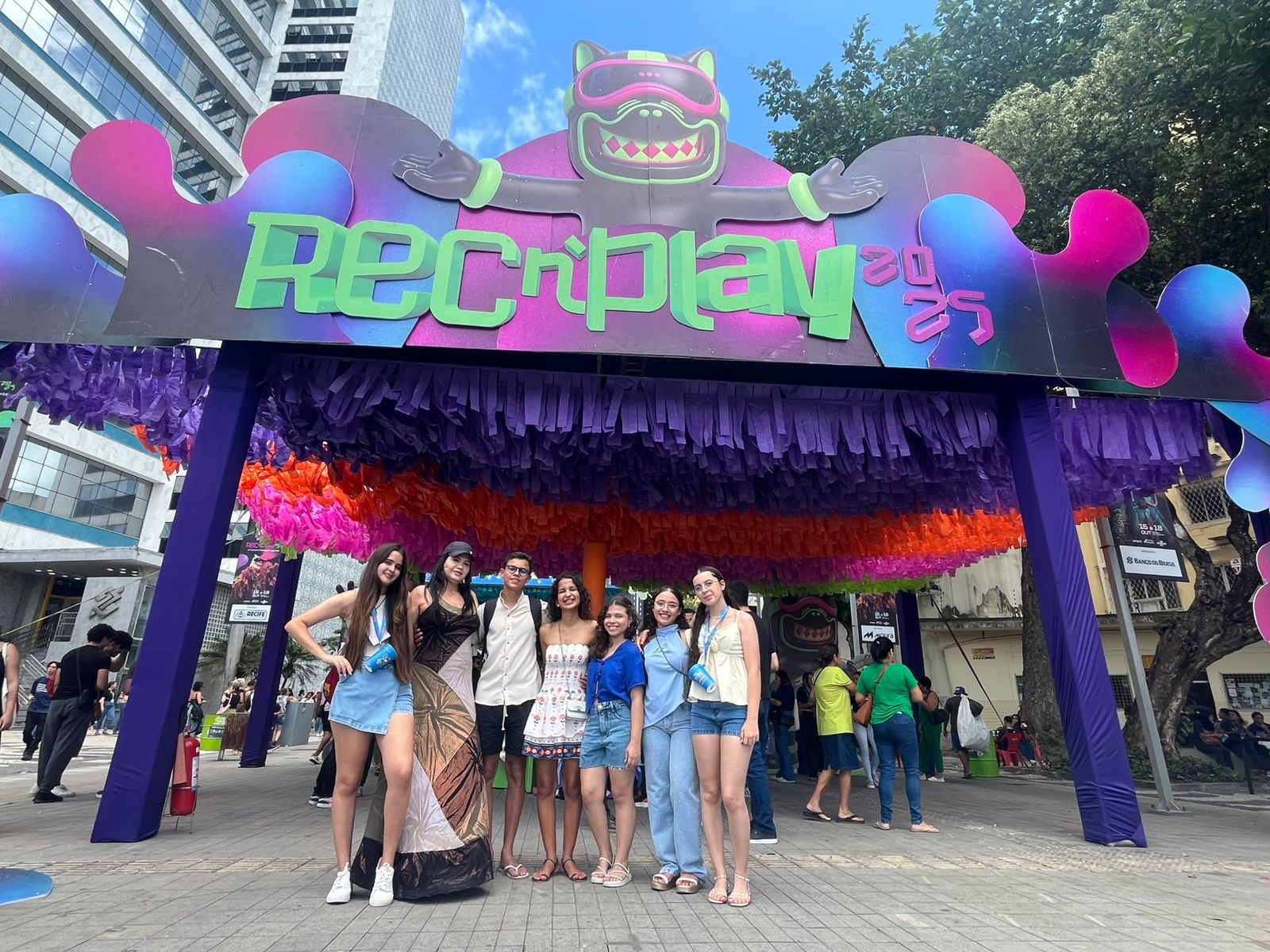 A professora Tatyana Keyty e seis estudantes do IFSertãoPE posam em frente ao pórtico principal de entrada do REC’n’Play 2025, no Bairro do Recife. O pórtico é grande, colorido e decorado com franjas de papel, exibindo a logo 'REC'n'Play 2025'