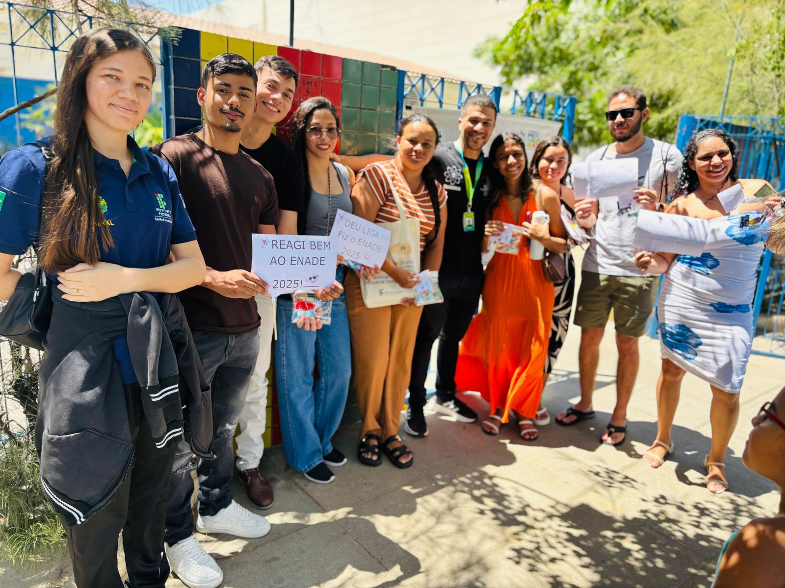 A foto mostra um grupo de aproximadamente dez pessoas, reunidas ao ar livre em um dia ensolarado, em frente a um muro colorido (vermelho, amarelo, verde e azul). No centro, há um homem de camisa preta e crachá, identificado como o professor Renato César, Coordenador do curso de Licenciatura em Química, ao lado de estudantes. Alguns estudantes seguram cartazes: Um estudante à esquerda segura um cartaz branco com a frase em português: "REAGI BEM AO ENADE 2025!". Uma estudante ao centro segura um cartaz ou sacola com a frase: "DEU LIGA !!! Rumo ao ENADE 2025". O grupo está sorrindo e olhando para a câmera. A cena representa o momento de apoio e incentivo do Coordenador e o espírito positivo dos discentes da Licenciatura em Química que participaram do Exame Nacional de Desempenho dos Estudantes (Enade).