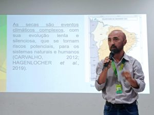 A imagem é uma fotografia do professor Washington Coriolano em pé, no canto inferior direito, apresentando uma palestra em um auditório.Palestrante: O professor, que é calvo e usa barba, veste uma camisa social de cor clara e segura um microfone com a mão direita, gesticulando com a mão esquerda. Ele usa um crachá de identificação pendurado no pescoço. Projeção (Slide): Ao fundo, um slide de apresentação em fundo claro está projetado. No lado esquerdo, em letras grandes na cor ciano, aparece a citação: "As secas são eventos climáticos complexos, com sua evolução lenta e silenciosa, que se tornam riscos potenciais, para os sistemas naturais e humanos (CARVALHO, 2012; HAGENLOCHER et al., 2019)." Contexto Geográfico: No lado direito do slide, há um mapa com uma demarcação geográfica, provavelmente sobre o semiárido brasileiro, relacionado ao tema da seca, risco e clima.