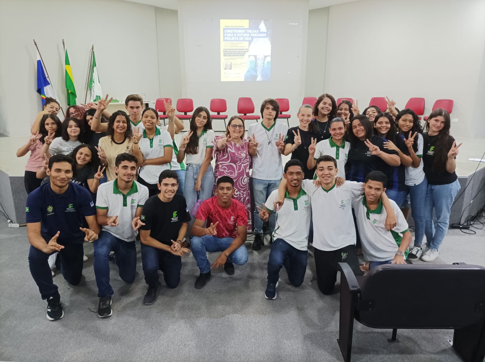 Foto em grupo no auditório do evento "Construindo Trilhas para o Futuro: Pensando Projeto de Vida". Um grupo entusiasmado de aproximadamente 30 pessoas, em sua maioria estudantes, está posando na frente das cadeiras do auditório. No centro do grupo, ligeiramente à esquerda, está a Psicóloga Lady Anne (a única adulta, vestindo uma roupa estampada em tons de rosa e roxo). Os estudantes estão sorrindo e fazendo o sinal de "paz e amor" com as mãos, muitos vestindo uniformes com as cores da instituição. Ao fundo, uma tela de projeção exibe o tema do evento. Bandeiras do Brasil e de Pernambuco estão visíveis no palco.