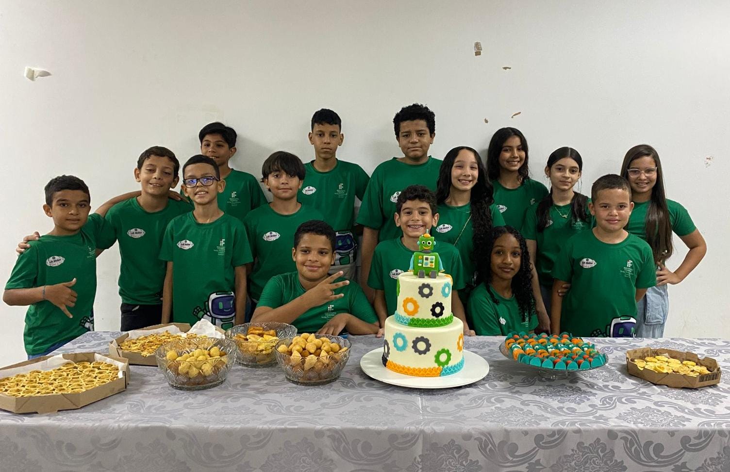 Foto em grupo celebrando o curso "LabMaker IF Ouricuri – Robótica e Inovação para o Sertão do Araripe". Quatorze crianças e pré-adolescentes, a maioria entre 10 e 12 anos, estão em pé atrás de uma mesa, olhando para a câmera e sorrindo. Todos usam camisetas verdes idênticas, que provavelmente são do curso. Na frente da mesa, que está coberta por uma toalha cinza, há diversas guloseimas: bandejas de salgados nas laterais, tigelas de doces ou salgadinhos no centro e, em destaque, um bolo de dois andares com decorações que imitam engrenagens coloridas (preto, laranja, verde) e um pequeno robô de brinquedo no topo. O ambiente é simples, com parede branca lisa. A pose indica uma celebração de encerramento ou de um marco do curso.
