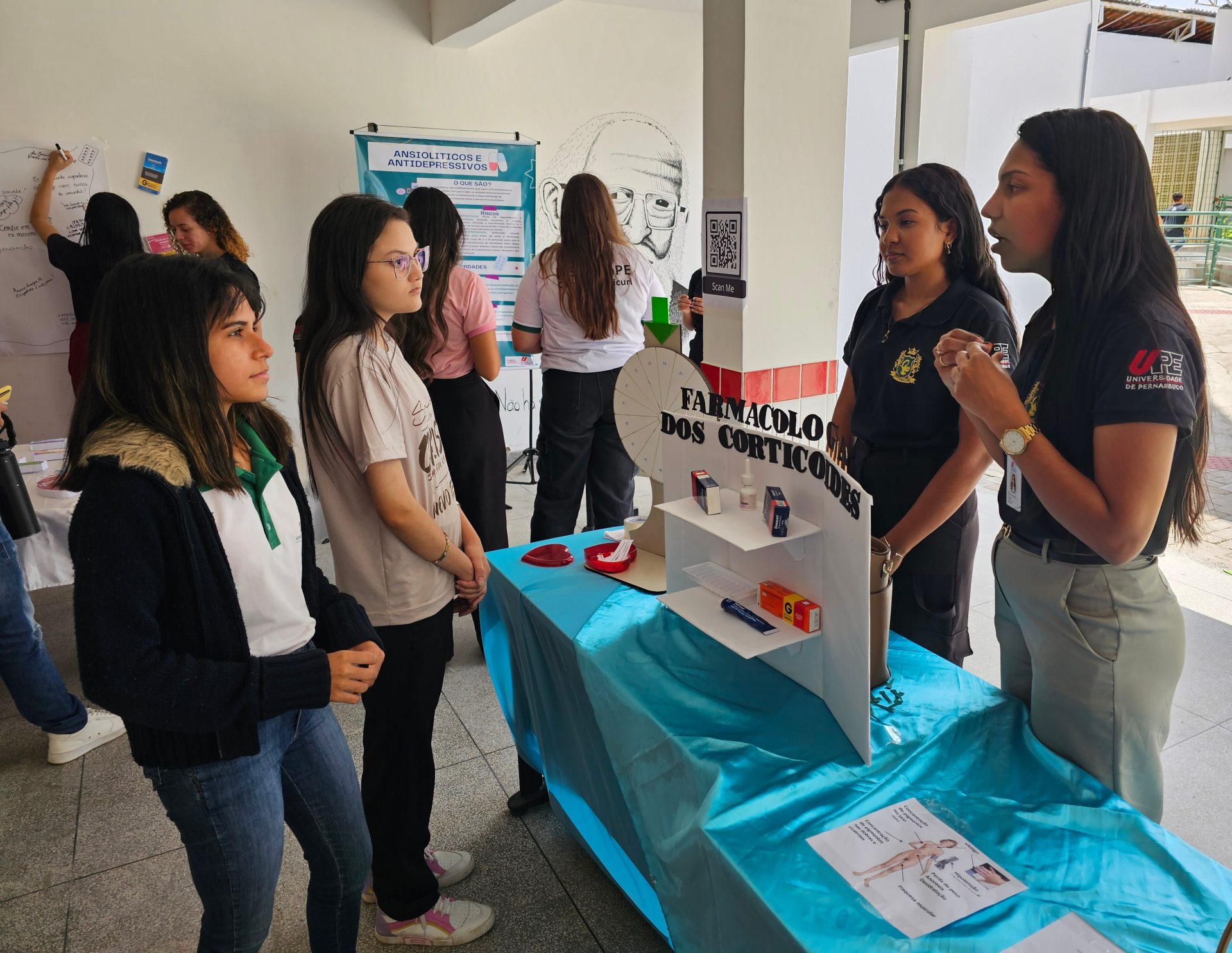 A imagem retrata uma Mostra de Farmacologia realizada pelos alunos do curso de Enfermagem do Campus Ouricuri da Universidade de Pernambuco (UPE), provavelmente nas dependências do Instituto Federal do Sertão Pernambucano (IFSertãoPE) - Campus Ouricuri, conforme a descrição. 💊 Detalhes da Cena e Conteúdo Tema Central: O estande em primeiro plano tem como tema a "FARMACOLOGIA DOS CORTICÓIDES" . O título está escrito em letras grandes em uma estrutura de painel com alguns modelos de medicamentos dispostos em prateleiras. Interação: Duas alunas/instrutoras, possivelmente do curso de Enfermagem da UPE, estão interagindo com duas visitantes/alunas, explicando o conteúdo do estande. Outros Temas: Ao fundo, é possível notar outro painel que trata de "ANSIOLÍTICOS E ANTIDEPRESSIVOS", indicando que a mostra aborda diversas classes de medicamentos e seus mecanismos de ação. Recursos Visuais: Há cartazes, displays e, no estande dos corticoides, um modelo que simula uma apresentação visual do tema, além de uma figura didática no chão que parece ilustrar o corpo humano e a ação dos fármacos. A mostra parece ser uma atividade de extensão ou didática para a comunidade acadêmica e/ou local, promovendo a disseminação de conhecimento essencial da farmacologia, uma disciplina fundamental para a área da saúde.