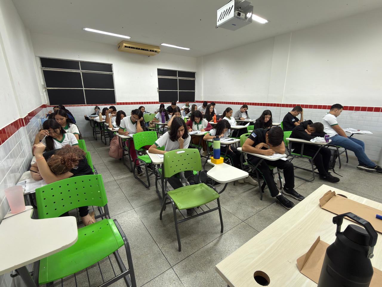 A foto, tirada de um ângulo ligeiramente elevado e lateral, mostra o interior de uma sala de aula escolar bem iluminada, onde um grupo de jovens (aparentemente adolescentes ou estudantes de ensino médio/universitário) está realizando algum tipo de teste ou exame. Ambiente: O ambiente é simples e funcional, com paredes brancas e uma faixa de azulejos vermelhos e brancos na altura da metade da parede. O piso é de azulejos claros. Há grandes janelas ou painéis escuros na parte superior da parede de fundo, possivelmente cobertos. Um projetor ou unidade de ar condicionado está fixado no teto. Mobiliário: Os estudantes estão sentados em carteiras individuais, com a maioria das cadeiras sendo de plástico em uma cor verde-limão vibrante. Alunos: Quase todos os estudantes estão com a cabeça baixa, concentrados em preencher ou escrever em papéis ou cadernos que estão sobre suas mesas. A maioria está vestindo camisetas brancas ou verdes e pretas. Há uma aluna em primeiro plano à esquerda que está descansando a cabeça no braço sobre a mesa, e um aluno na frente e no centro que parece estar lendo atentamente o seu material. Primeiro Plano: No canto inferior direito, há uma mesa maior (possivelmente a do professor) onde se vê uma garrafa térmica preta e outros objetos. Em resumo: É uma cena de estudo ou avaliação formal em um ambiente escolar interno.