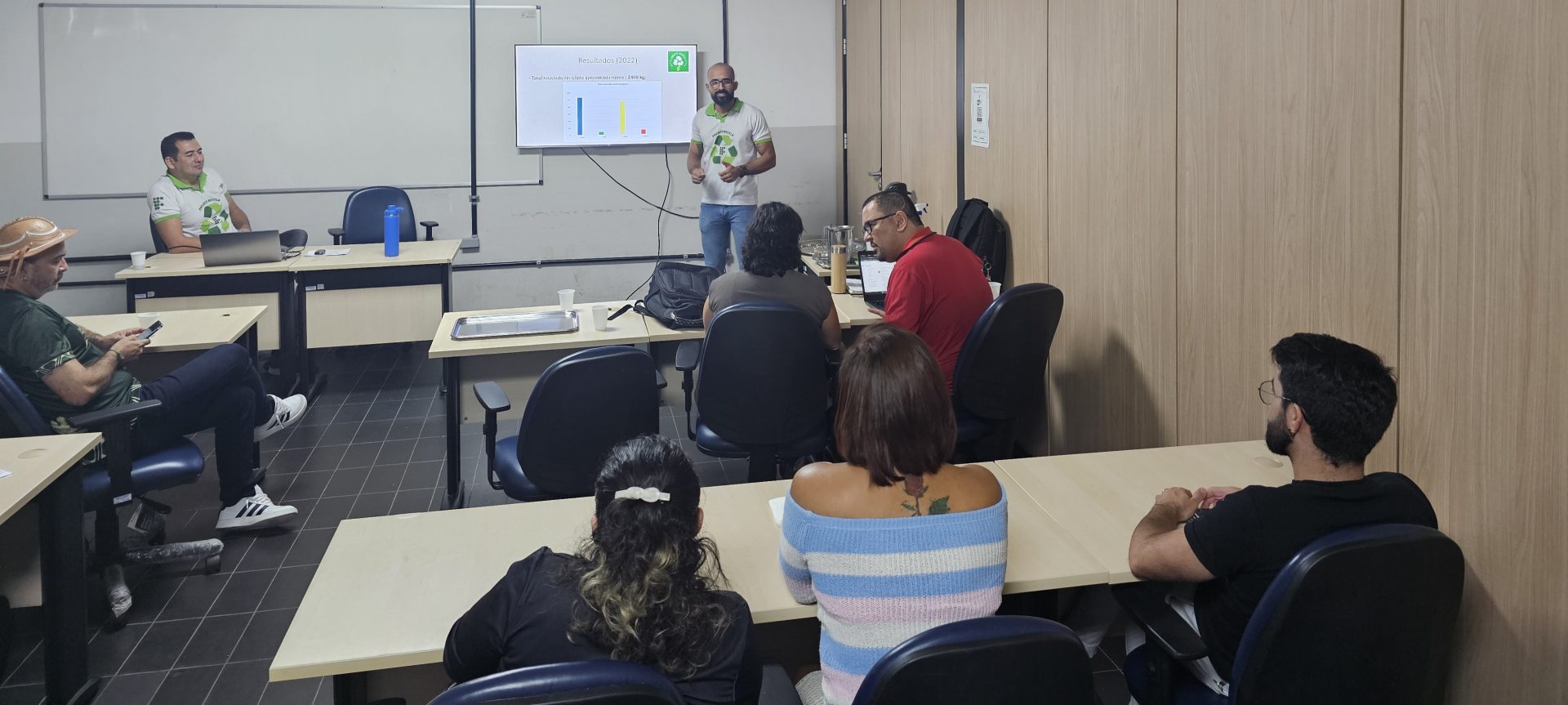 Uma palestra está sendo ministrada em uma sala de aula. Um homem com barba, camiseta branca com estampa verde e óculos escuros está de pé na frente da sala, apontando para uma tela branca onde gráficos são exibidos. Há mesas e cadeiras com várias pessoas sentadas e atentas. Algumas pessoas usam camisetas semelhantes à do palestrante, com a mesma estampa verde.