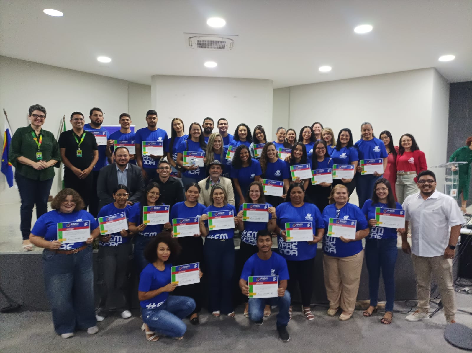 É uma fotografia em ambiente interno, em um palco ou área de apresentação, iluminada. Um grande grupo de pessoas, parecendo ser cerca de 40 a 50, está posicionado para uma foto. A maioria das pessoas está vestindo camisetas azuis vivas com a palavra "EMPREENDA" impressa em branco na frente, ou possivelmente outra palavra relacionada, e segurando certificados ou diplomas. Há várias fileiras de pessoas, algumas em pé e outras sentadas ou agachadas. Na frente, há pessoas sentadas ou agachadas, principalmente com camisetas azuis, segurando seus certificados. Uma mulher no canto inferior esquerdo está usando uma calça jeans azul-escura e a camiseta azul, enquanto um homem no canto inferior direito também está agachado. No meio e atrás, o grupo está em pé. Muitos parecem estar felizes e sorrindo. Algumas pessoas se destacam na vestimenta: Duas pessoas no canto superior esquerdo (um homem e uma mulher) estão com camisas mais escuras (verde-escura e preta, respectivamente) e cordões de identificação. Três homens no centro do grupo, ligeiramente à frente, estão vestindo camisas ou blazers mais formais (um com terno escuro, outro com camisa social clara e chapéu, e outro com camisa escura). Uma mulher na extrema direita, no fundo, está usando uma blusa vermelha vibrante e calças claras. Um homem na extrema direita está com uma camisa branca e calças claras. O fundo é uma parede bege clara ou cinza, com um canto mais escuro à direita. O palco tem um piso escuro. Há um ar-condicionado aparente na parede de fundo. A iluminação é brilhante, vinda de cima. A cena sugere uma cerimônia de formatura ou conclusão de curso/programa, provavelmente relacionado a "empreendedorismo" (dado o texto nas camisetas).