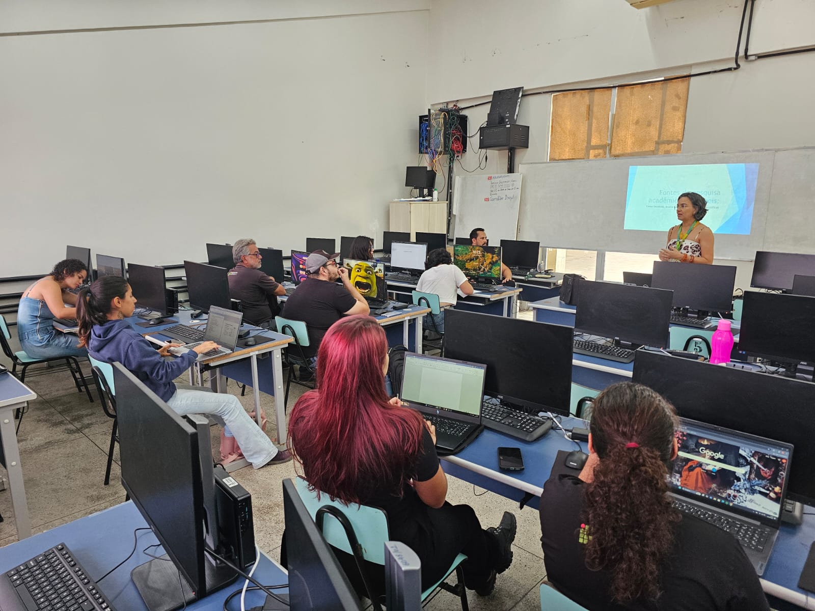 A imagem mostra uma sala de aula de informática, com vários computadores de mesa distribuídos em fileiras. Alguns estudantes estão utilizando notebooks próprios, enquanto outros parecem acompanhar o conteúdo projetado na tela ao fundo. Uma mulher, em pé ao lado do quadro branco, conduz a atividade. Na projeção, é possível ler parte do título “Fontes de pesquisa acadêmica...”. Os participantes estão atentos, e o ambiente transmite a ideia de uma oficina ou curso de capacitação.